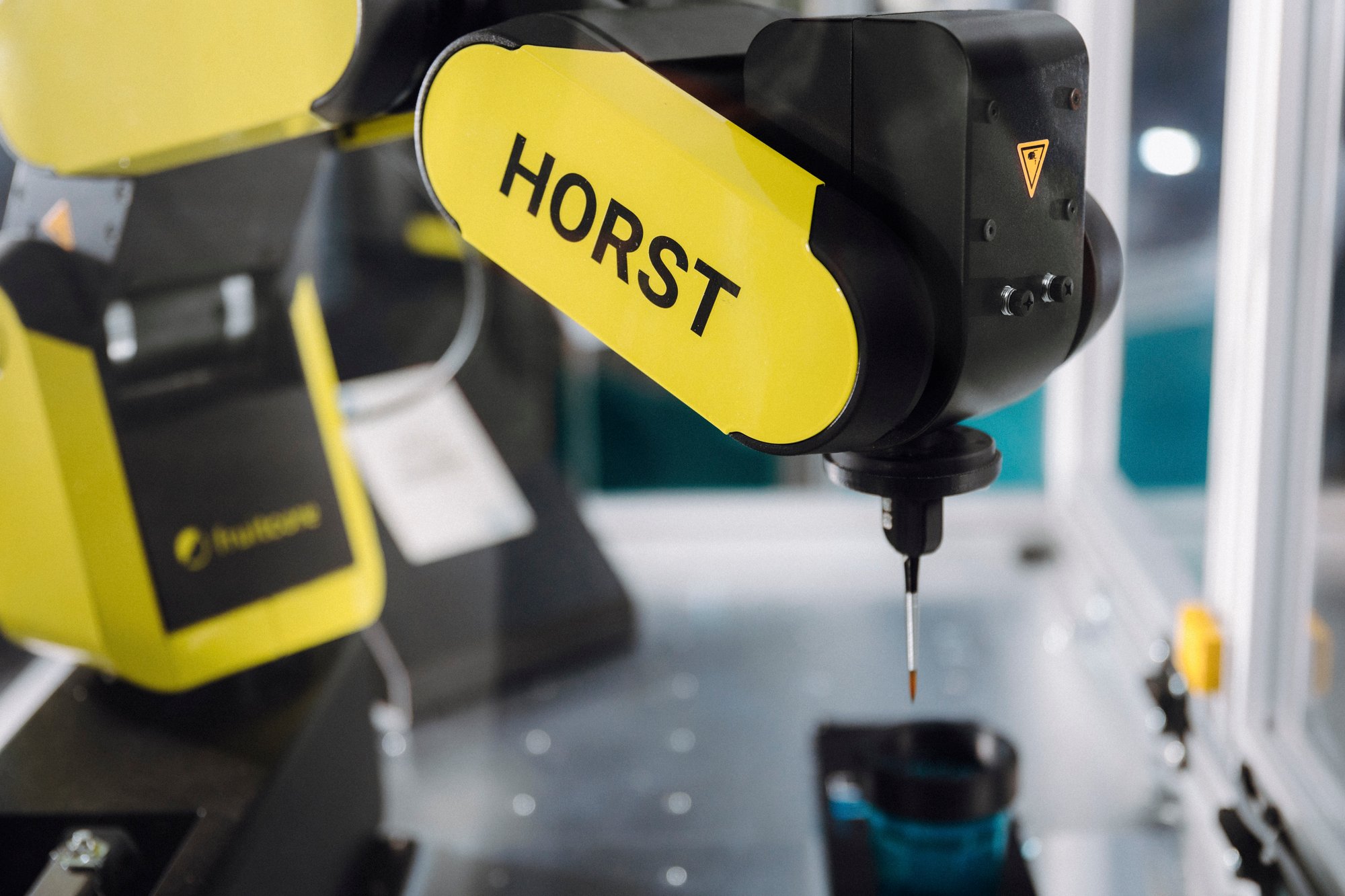 A yellow Horst600 robot arm with an attached paintbrush and a glass of water underneath