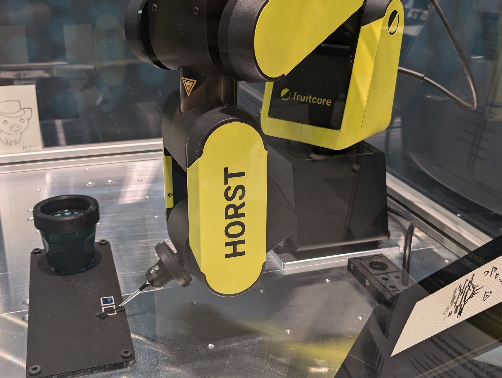 A yellow Horst600 robotic arm with an attached paintbrush, a glass of water and paint 